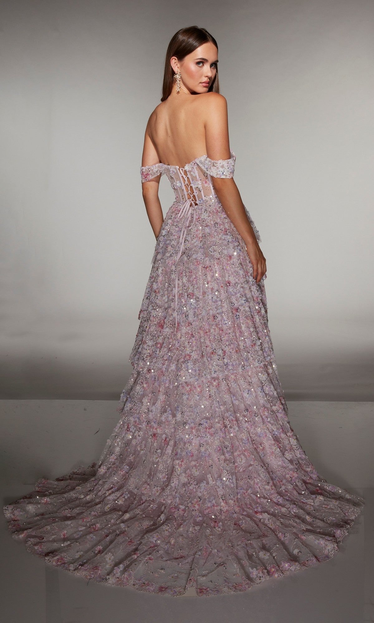 A woman poses with her back to the camera in the Alyce 62186 long prom dress, showcasing its lace-up back, pastel pink and purple a-line skirt, off-the-shoulder neckline, 3D floral appliques, and layered train.